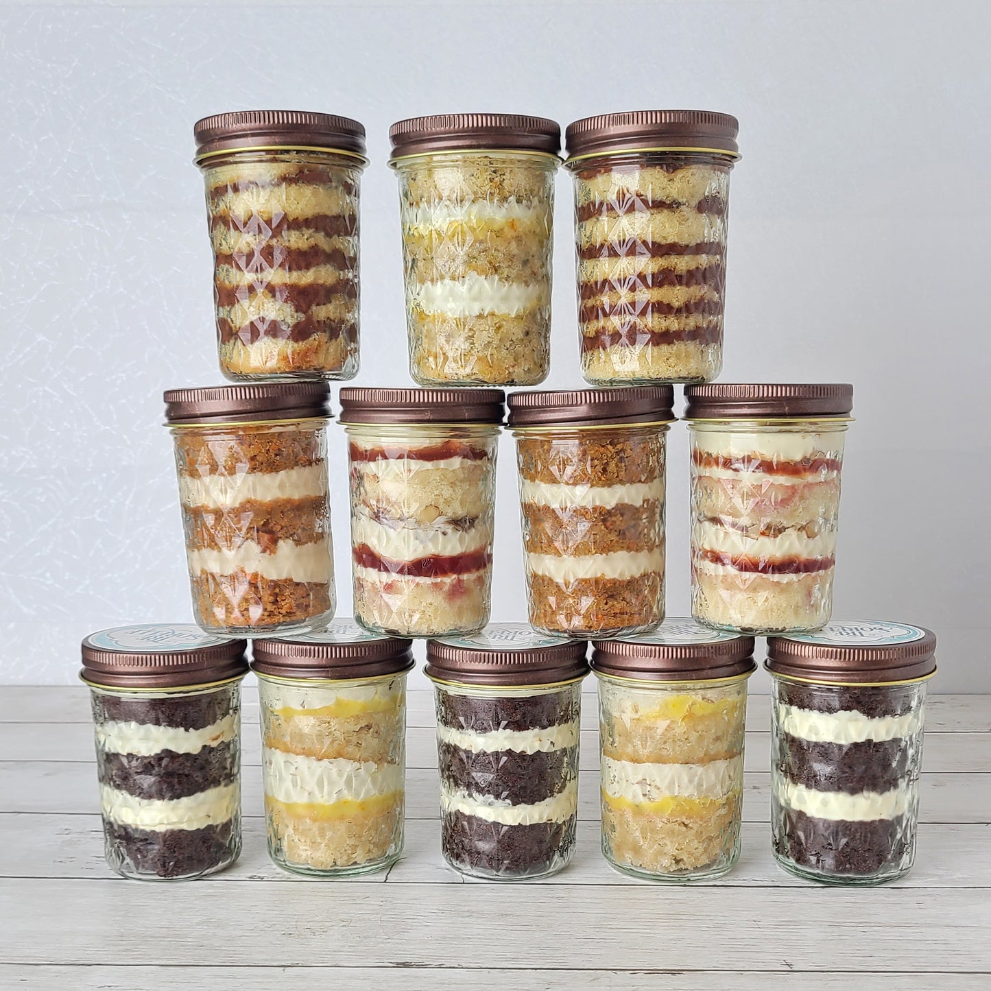 A pyramid of twelve fancy faceted glass jars filled with layers of cake and buttercream of various flavors. The jars are sealed with rustic bronze colored lids.