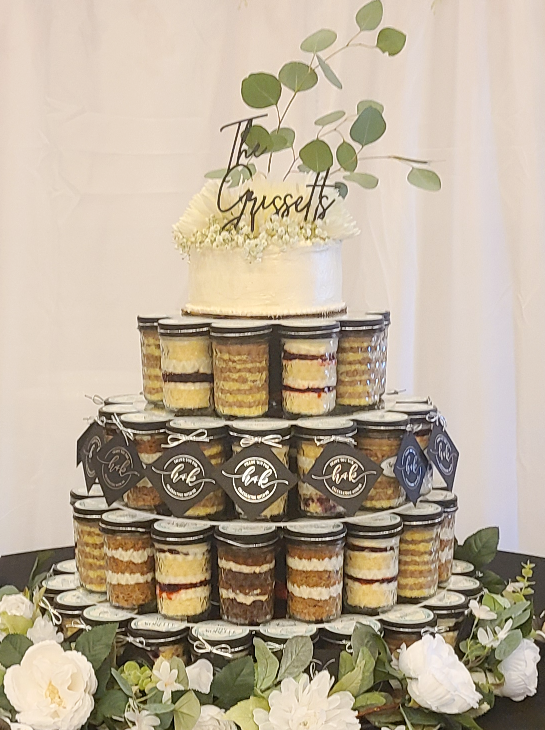 Individual cake-in-a-jar desserts stacked tall in concentric circles topped with a round cutting cake and adorned with florals and leaves. The setup is surrounded by white flowers and greenery, with a soft, elegant ambiance.