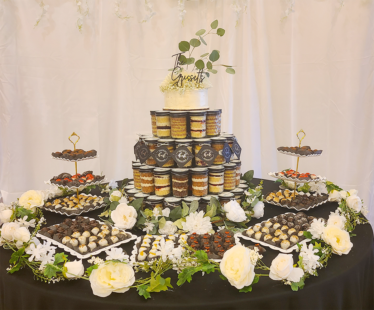 A wedding dessert table features a tiered wedding cake made up of many individual cake-in-a-jar favors.  Surrounding the display are trays of assorted cake truffles. White flowers and greenery on a black tablecloth add elegance.