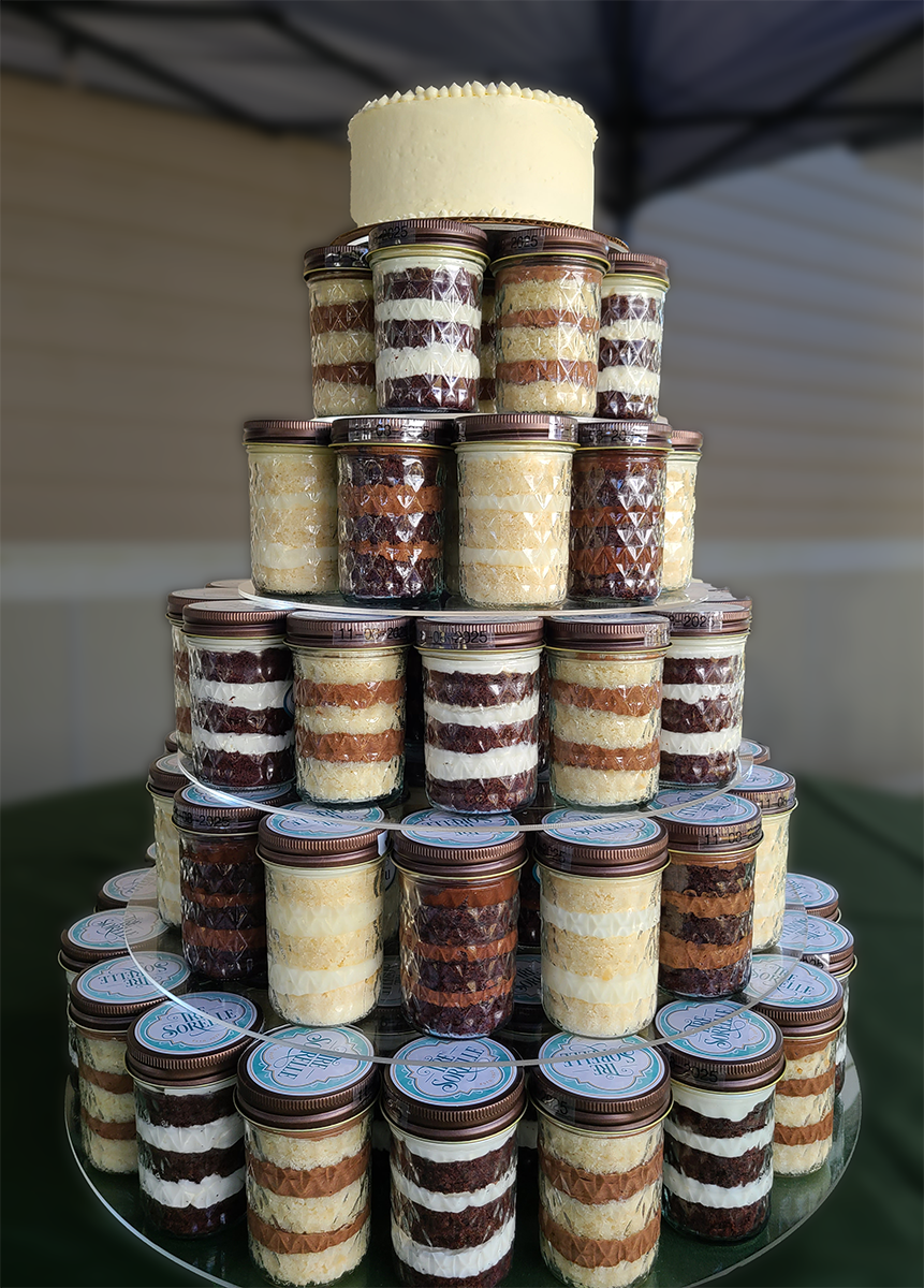 A tiered wedding cake display featuring cake-in-a-jar in assorted chocolate and vanilla combinations. Atop the display sits a small, frosted round cake.