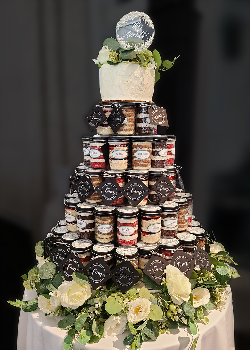 Three-tiered wedding cake made of stacked cake-in-a-jar with labels and favor tags, surrounded by flowers on a dark background.